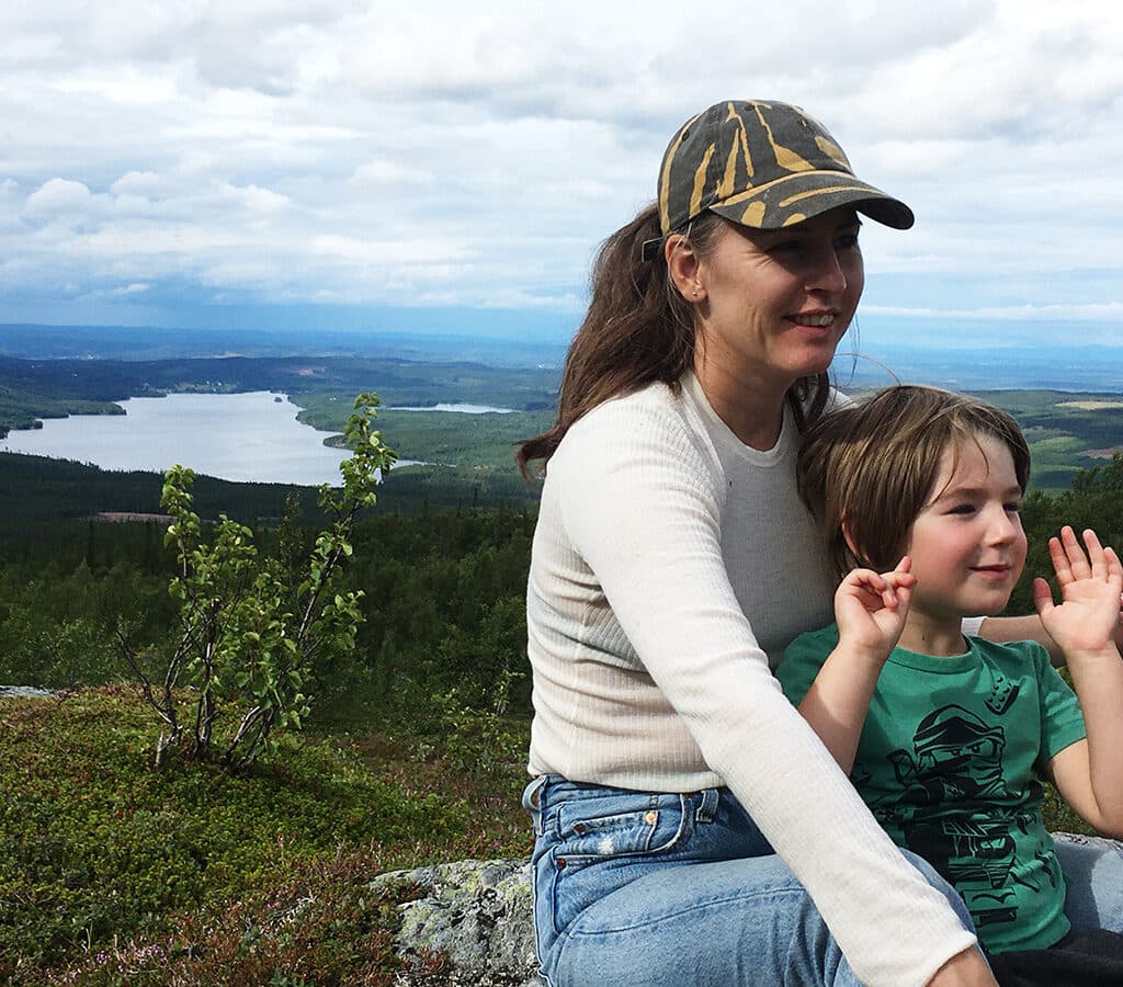 En person med keps och hästsvans sitter med ett barn mellan sina barn. De är glada och barnet vinkar med händerna