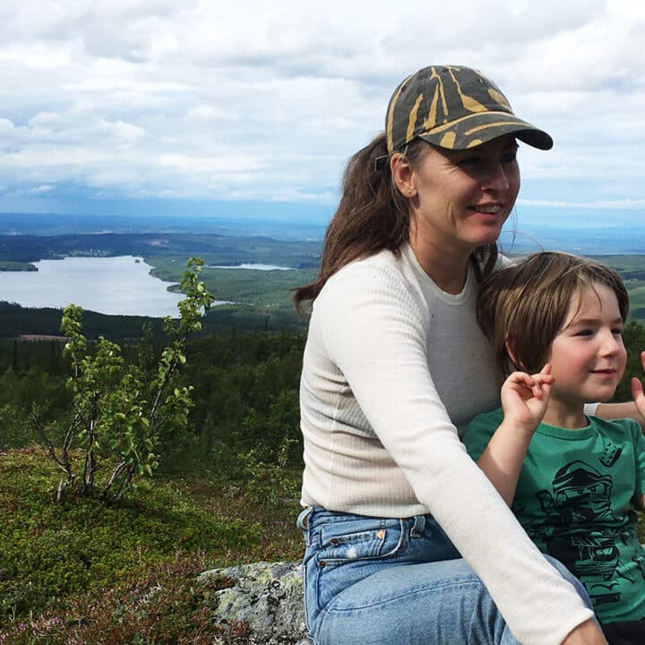 En person med keps och hästsvans sitter med ett barn mellan sina barn. De är glada och barnet vinkar med händerna